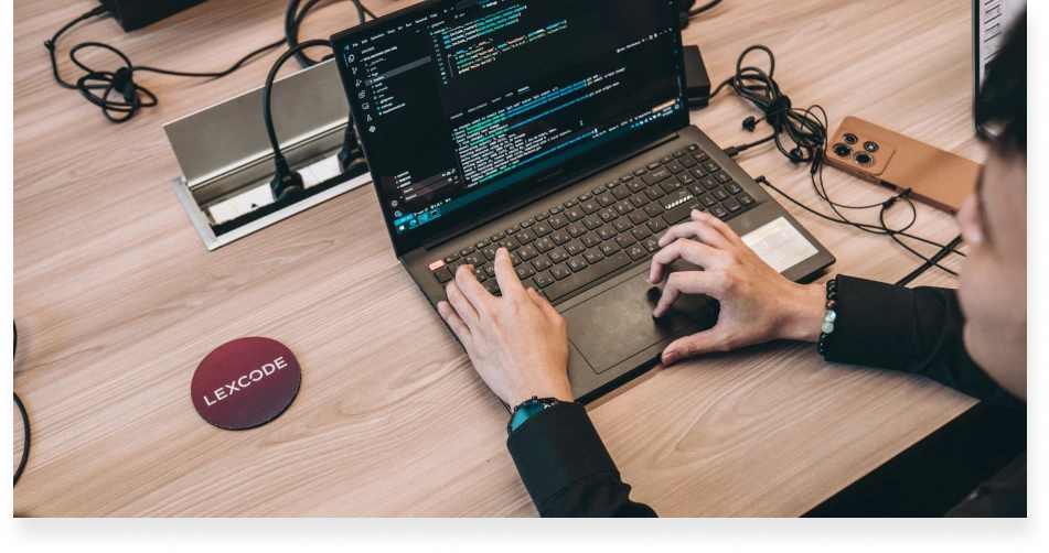 A software developer coding on a laptop in a Lexcode workspace, with a branded coaster beside the laptop.