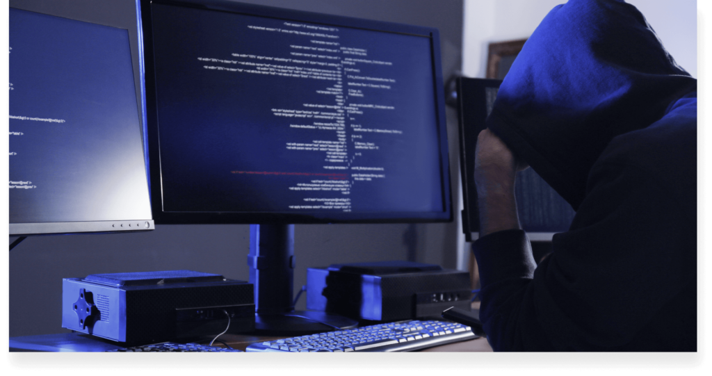 A person in a hooded jacket reviews code on a computer screen with a dark ambiance.