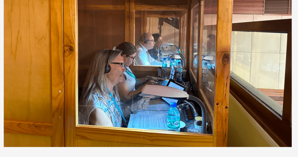 Four people, sitting side-by-side inside 2 interpretation booths with recording equipment and other materials arranged on the table.