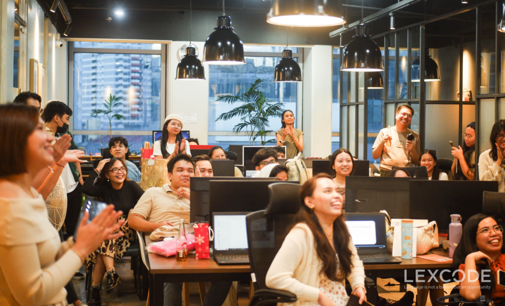 Various people standing and sitting on office chairs gather around to watch a Christmas video montage being played at the front of the room (unseen).