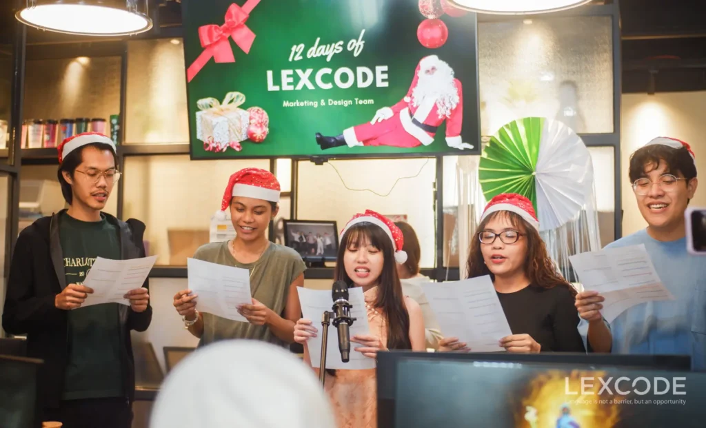 A group of men and women wearing Christmas hats sing with lyrics in hand at the front.