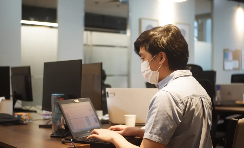 A person wearing a face mask working on a laptop in an office environment.