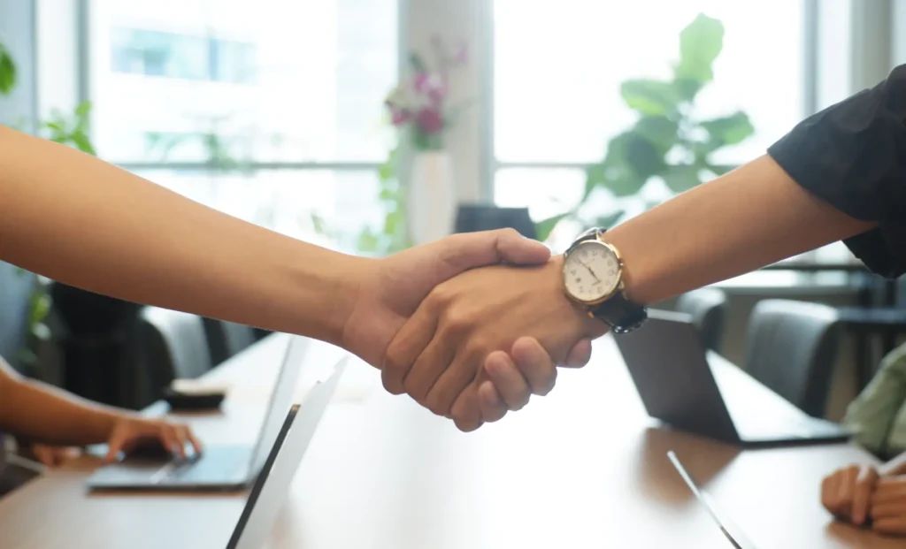 Two individuals shaking hands over a table with laptops in a meeting room.