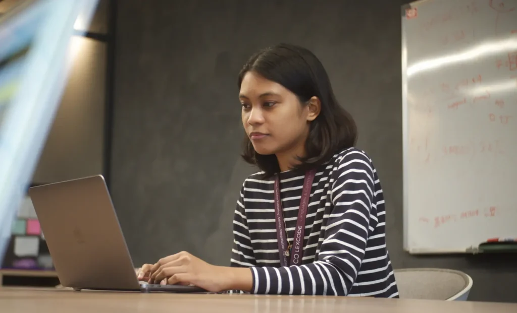 A young woman in a striped shirt typing on a laptop in a modern office space.