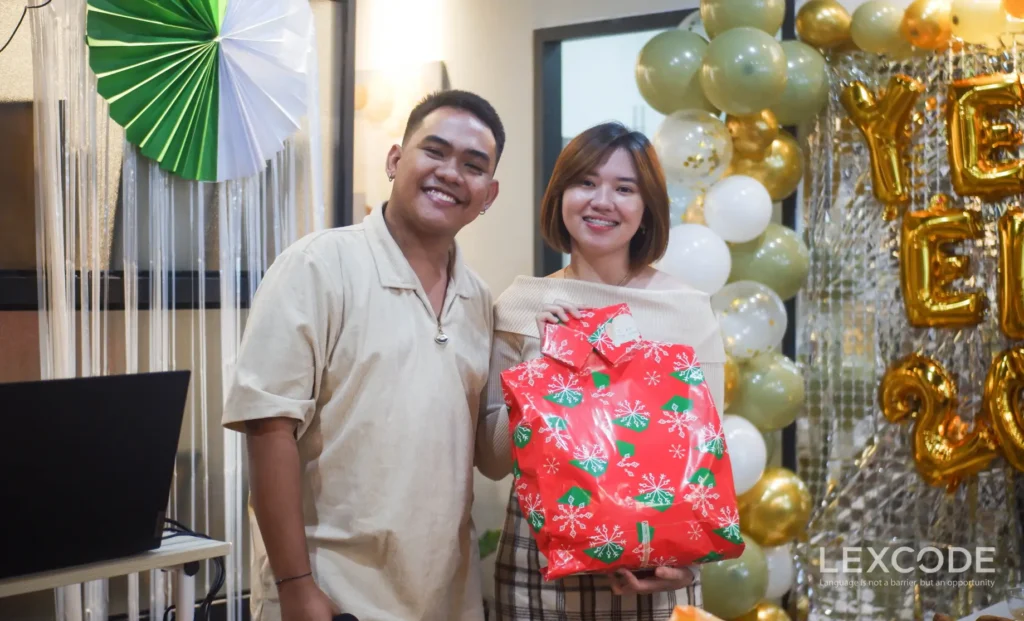 A man and a woman are smiling at the camera with the other person holding her Christmas gift.