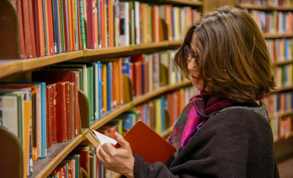 An anonymous woman sitting in a city library, immersed in reading a book, surrounded by bookshelves and warm ambient light.