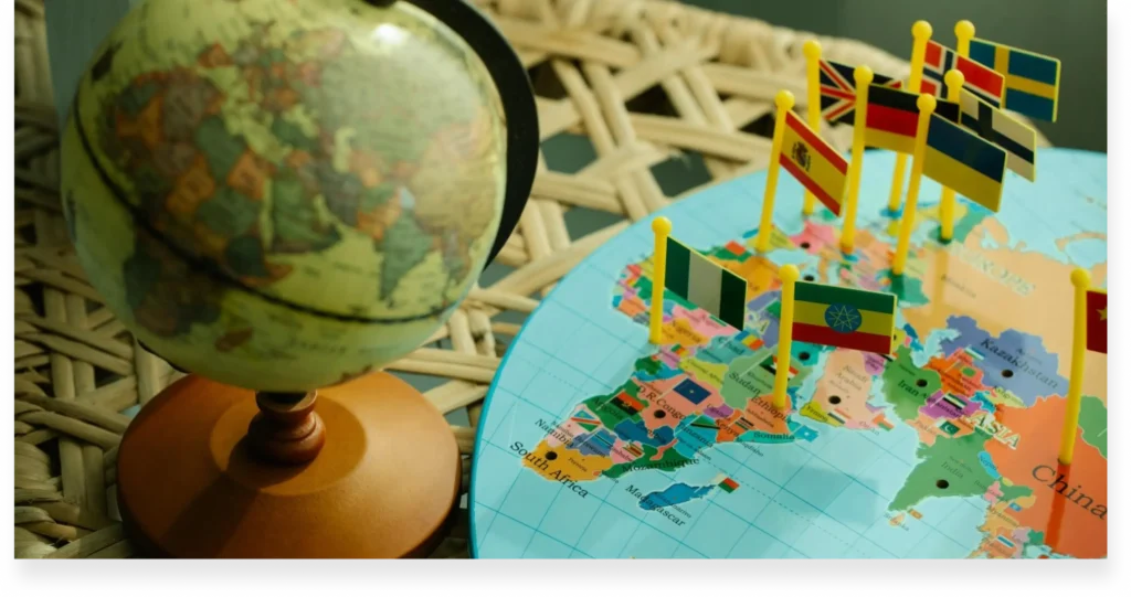 A world globe beside a map with various national flags marking countries on a wicker surface.