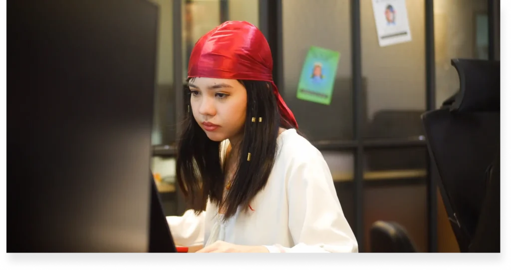 A woman in white shirt and red headscarf seated at a desk with monitors, office setting in the background.