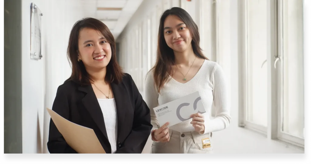 Two (2) women posing outside of an office, holding official documents and an envelope.