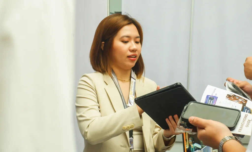 The former employee demonstrating an AI-powered product to an attendee holding a flyer and mobile phone.