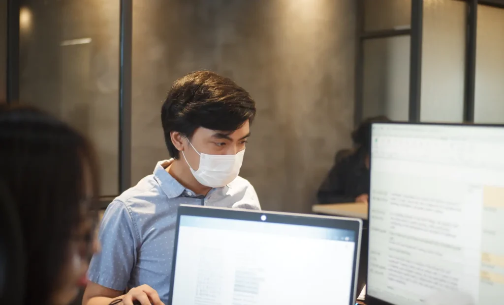 A person wearing a face mask working on a laptop in an office environment.