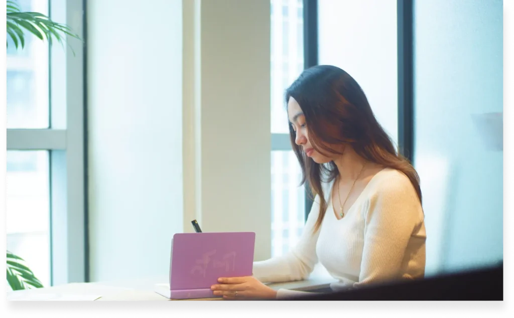 Woman writing in a notebook while seated by a large window in a modern office.
