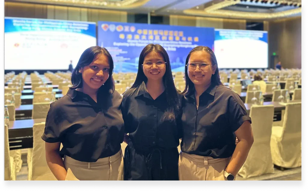 Jeniña, Bea, and Karla smiling together at an eQQui event, with a conference hall setup in the background.