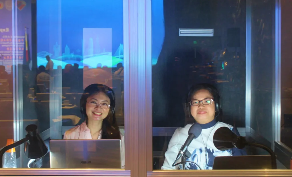 Two interpreters inside a booth during a live event, wearing headsets and using laptops.