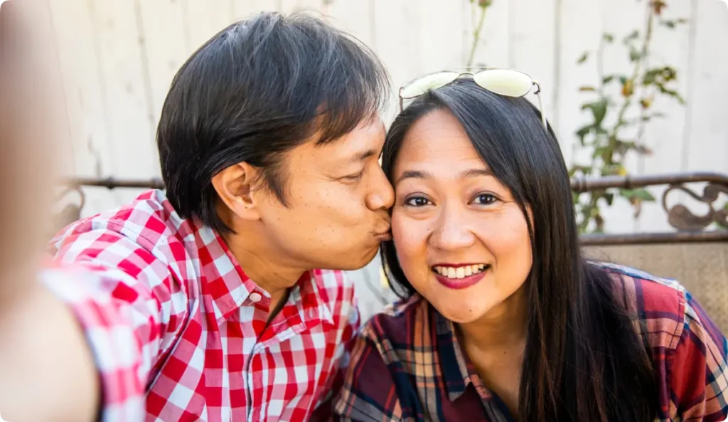 Happy Asian couple taking selfie with man kissing woman's cheek on sunny balcony