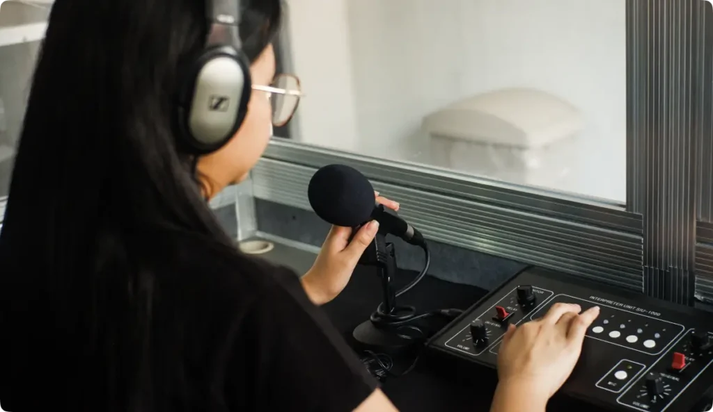 Professional female interpreter in translation booth wearing headset and using microphone with control panel