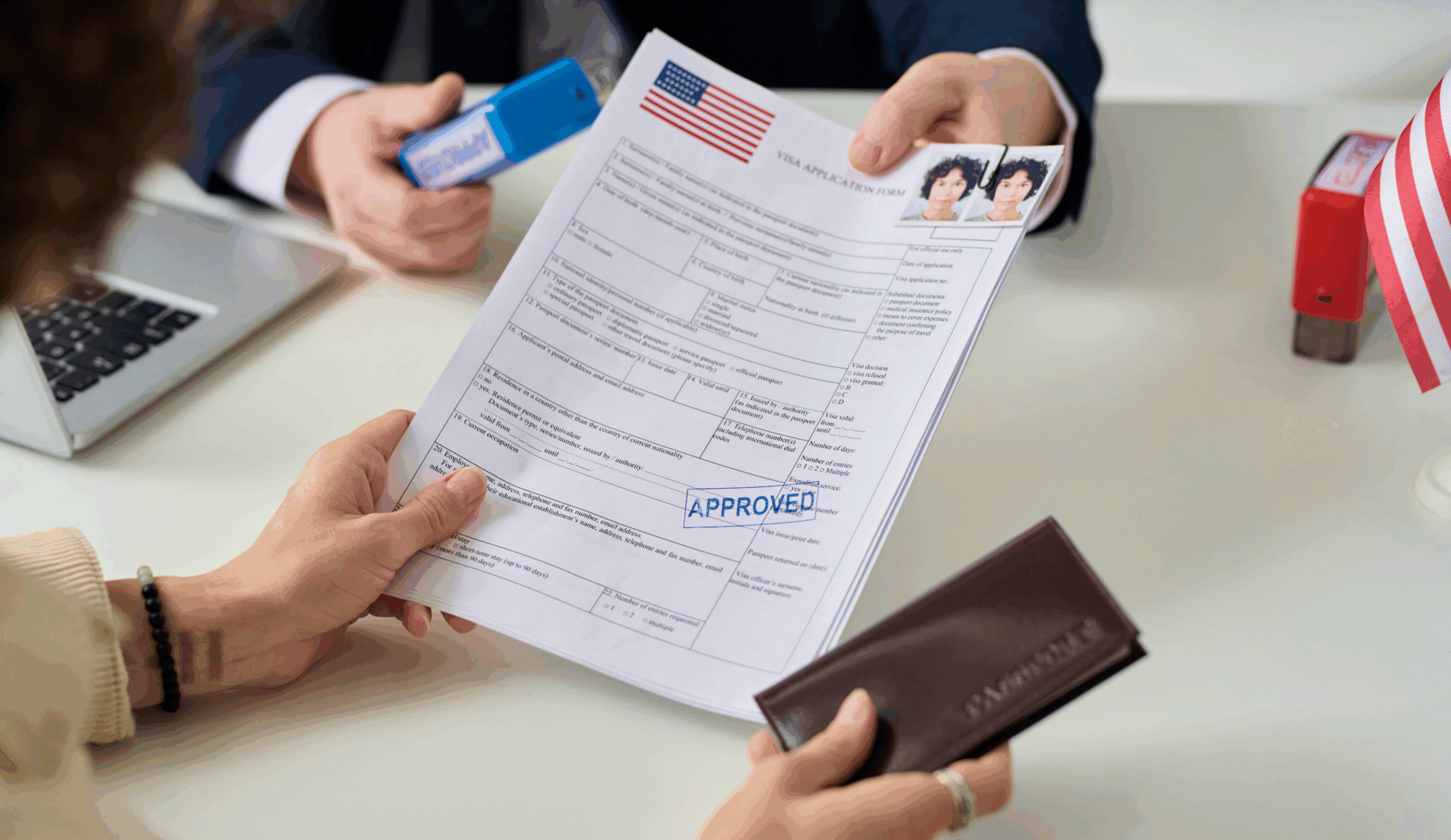 A person holding an approved visa document at a table while another person uses a blue stamp, with passports and a laptop nearby.